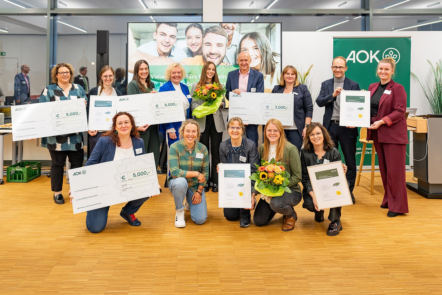 Das Foto zeigt die Preisträger des GesundMacher Awards. Reihe vorne (v.l.n.r.): Verena Neubauer (Universitätsklinikum Erlangen), Laura Dahlmeier, Elke Schmidt und Bianca Adler (Universitätsklinikum Erlangen), Ulrike Nöth (Gesundheitsnetz QuE) Reihe hinten (v.l.n.r.): Katharina Ehinger und Jasmin Hildner (Diakonisches Werk Coburg), Alina Wimmer und Cornelia Uschold (Constantia Pirk), Julia Oelmayer (Gesundheitsnetz QuE), Horst Leitner (Direktor AOK Nürnberg), Annette Lutz (Bereichsleitung Gesundheitsförderung AOK Bayern), Stephan Abele (Vorstandsvorsitzender der AOK Bayern), Christina Heinemann (Universitätsklinikum Erlangen)