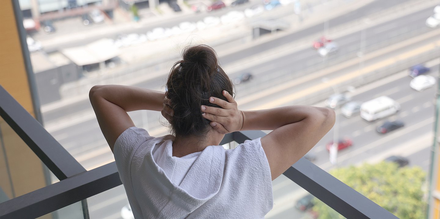 Foto: Eine Frau steht auf einem Balkon, unter ihr fahren Autos. Sie hält sich die Ohren zu.