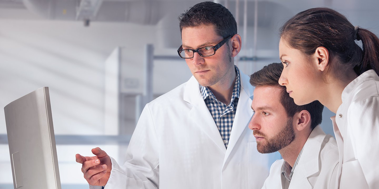 Ein Besprechungsraumim Krankenhaus. Ein Team aus Ärzten schaut gemeinsam auf den Bildschirm. Dabei zeigt ein Arzt auf etwas, was auf dem Monitor dargestellt wird.