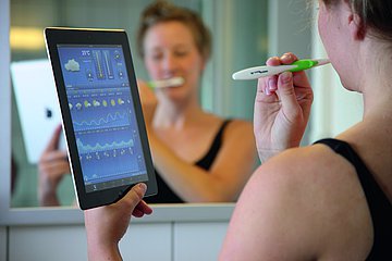 Gesund mit App und Web Eine Jugendliche schaut selbst beim Zähneputzen auf ihr Tablet.