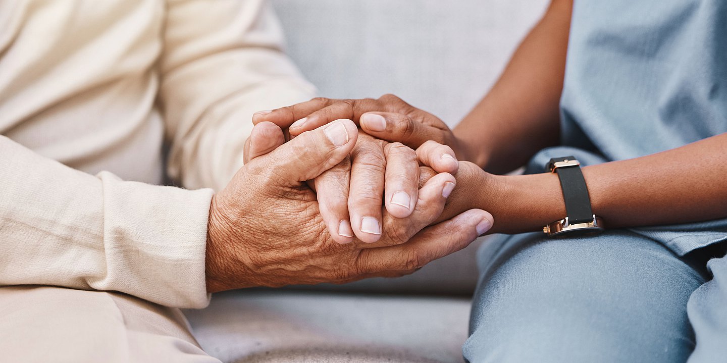 Foto: Die Hand einer jüngeren Frau in Pflegetracht hält die Hand einer älteren Frau.