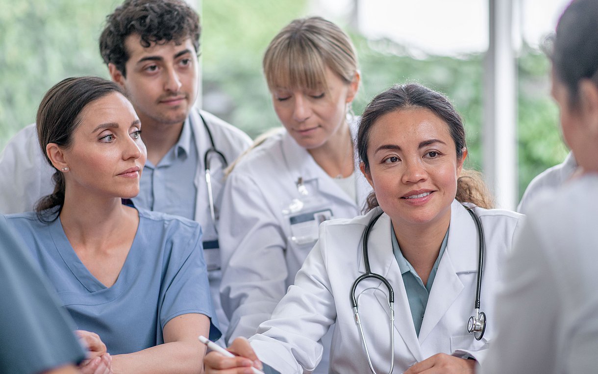 Das Foto zeigt eine Gruppe von sechs Medizinern (fünf Frauen, ein Mann) im Gespräch offebsichtlich um einen Stehtisch herum, zwei der Frauen wenden dem Betrachter den Rücken zu.