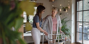 Doppelbelastung von Frauen Foto einer jungen Frau, die in einem Wohnzimmer einer alten Dame am Rollator hilft