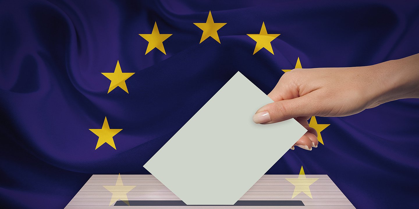 Eine Hand steckt einen Briefumschlag in eine Wahlurne vor einer Europaflagge.