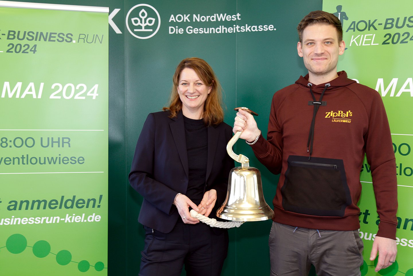 Das Foto zeigt AOK-Landesdirektorin Iris Kröner und Geschäftsführer Paul Ziplinsky von ZIPPEL'S Läuferwelt mit einer großen Glocke zur Vorankündigung des AOK-Business.Run Kiel. eine große Glocke in der Hand, die von Iris Krönehält