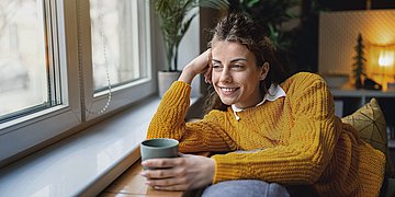 Woche der seelischen Gesundheit Foto einer jungen Frau, die sich glücklich grinsend mit einer Tasse Tee in der Hand auf ein Fensterbrett stützt und nach draußen blickt