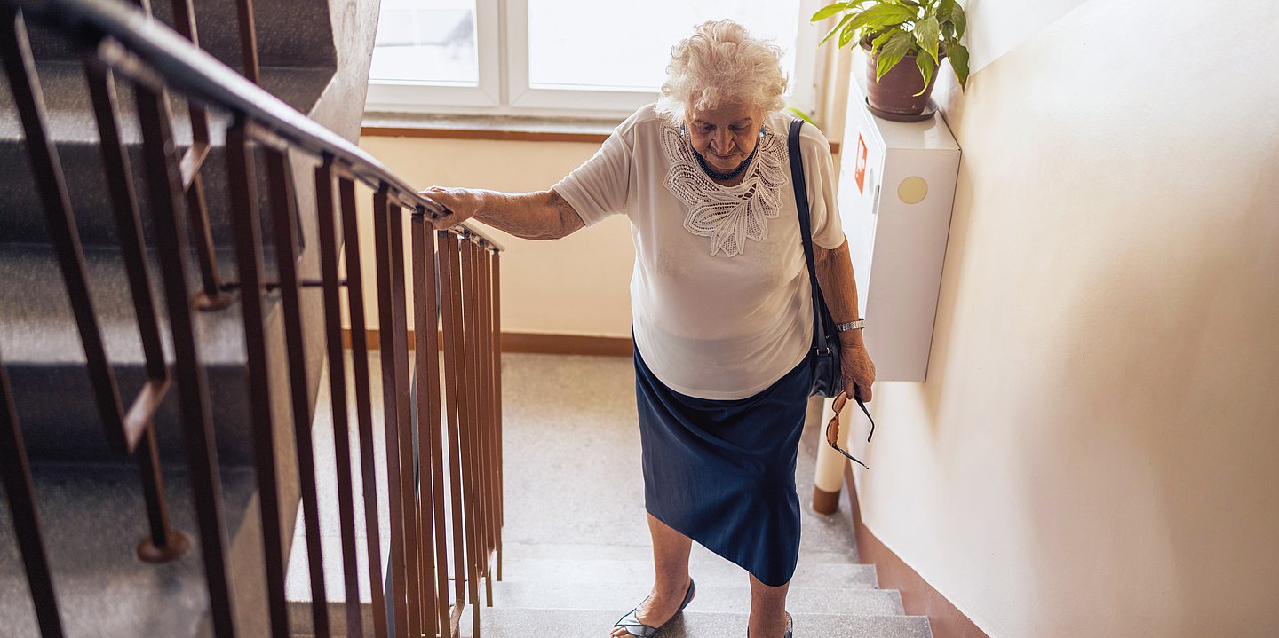 Foto: Eine ältere Dame, läuft Treppen, hält sich am Geländer fest und sieht angestrengt aus.