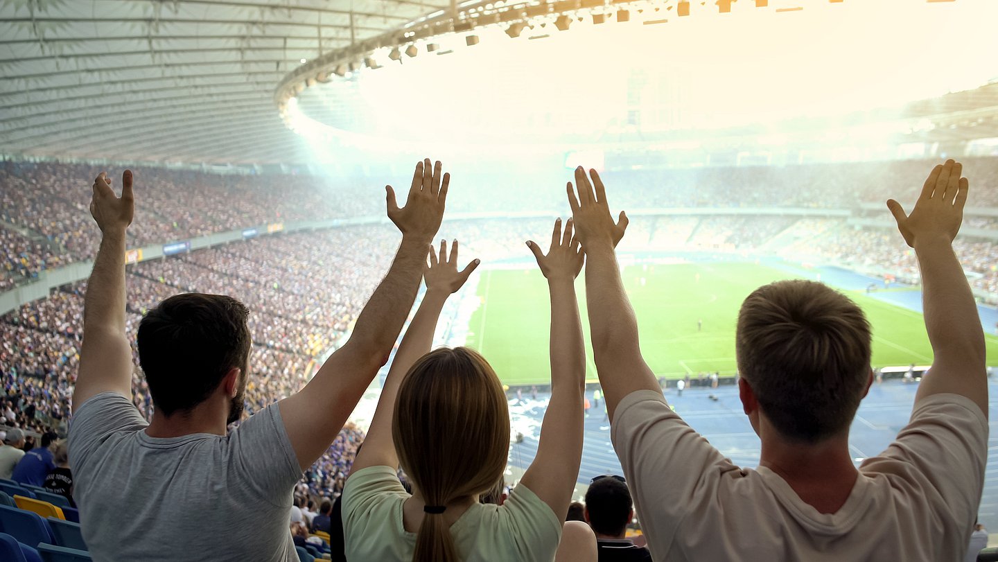 Foto zeigt ein volles Fußballstadion mit Blick auf das Spielfeld und jubelnde Fans, die die Arme hochreißen.