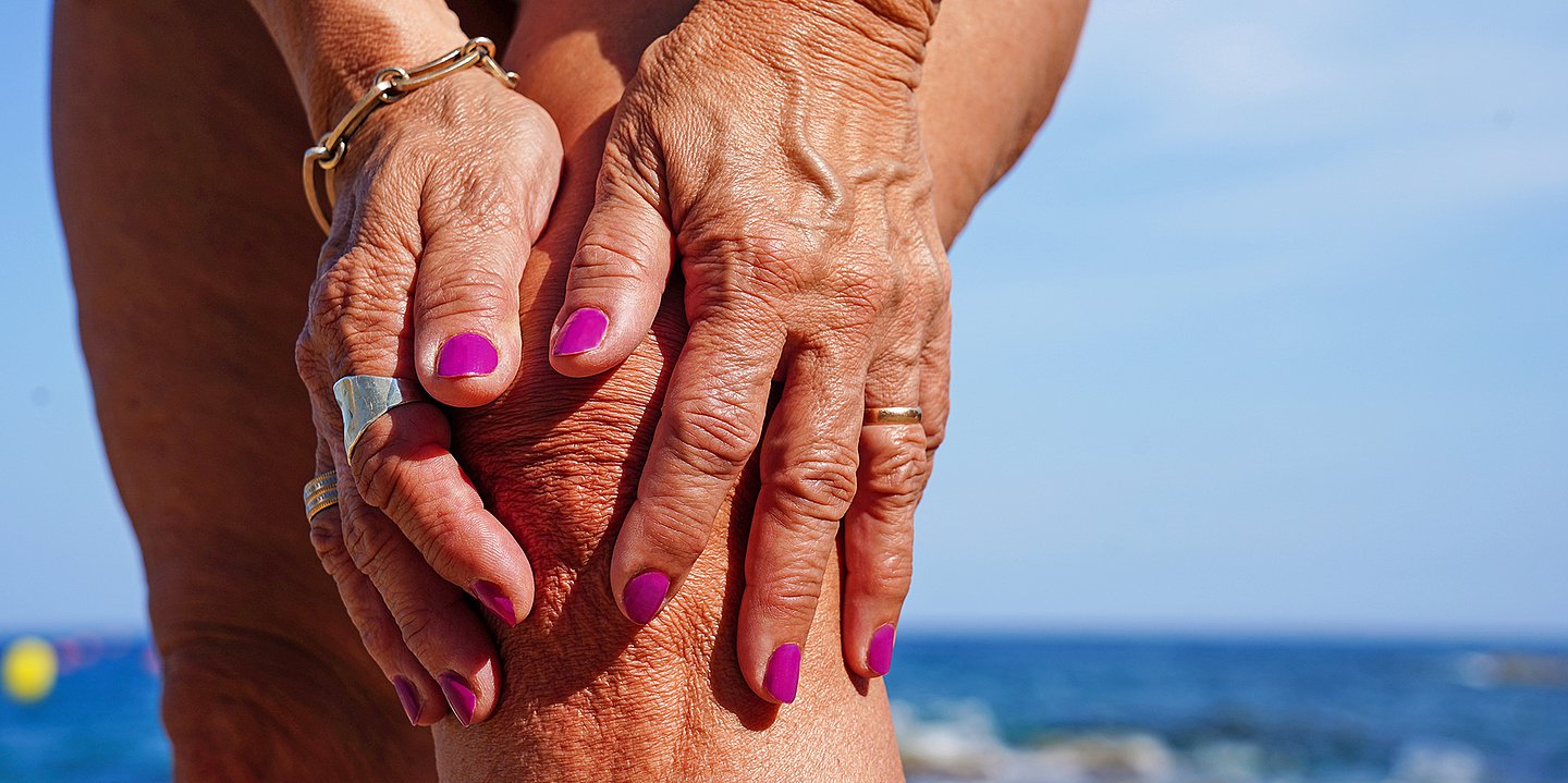 Foto einer Frau am Strand, die sich das Knie hält. Es sind nur Teile ihrer Beine und ihre Hände mit Nagellack zu sehen.