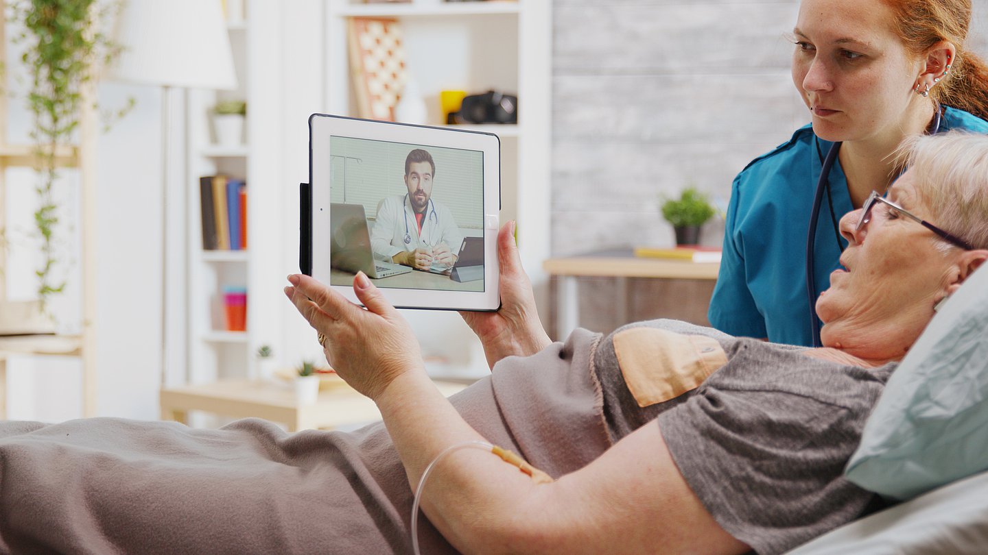 eine Patientin, die zu Hause oder im Pflegeheim lebt, führt mit Hilfe einer Pflegerin per  Videosprechstunde ein Gespräch mit ihrem Arzt. Dabei kann sie bequem auf dem Sofa liegen, während sie den Bildschirm hält.