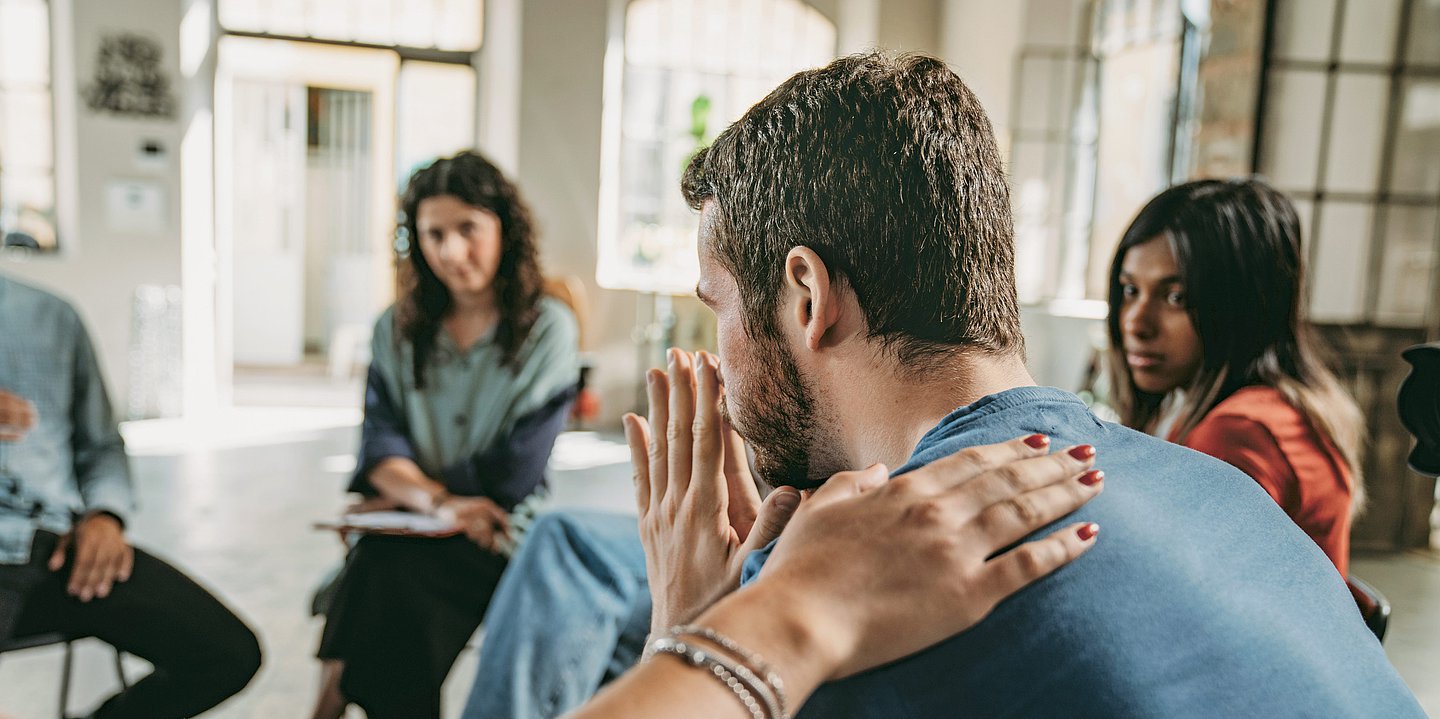 Foto: Einige Menschen sitzen in einem Stuhlkreis, ein Mann hält sich die Hände vor das Gesicht, während eine andere Hand tröstend auf seinem Rücken liegt.