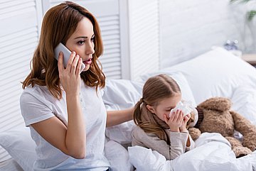 Kinderkrankengeld Schleswig-Holstein Foto zeigt eine Mutter, die am Bett ihres kranken Kindes sitzt und ein Telefongespräch führt.