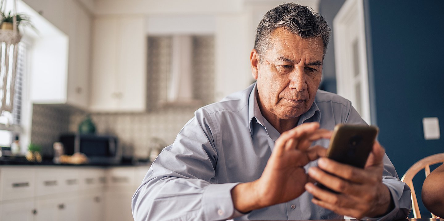 Das Bild zeigt einen älteren Herrn mit graumeliertem Haar und Smartphone am Küchentisch.