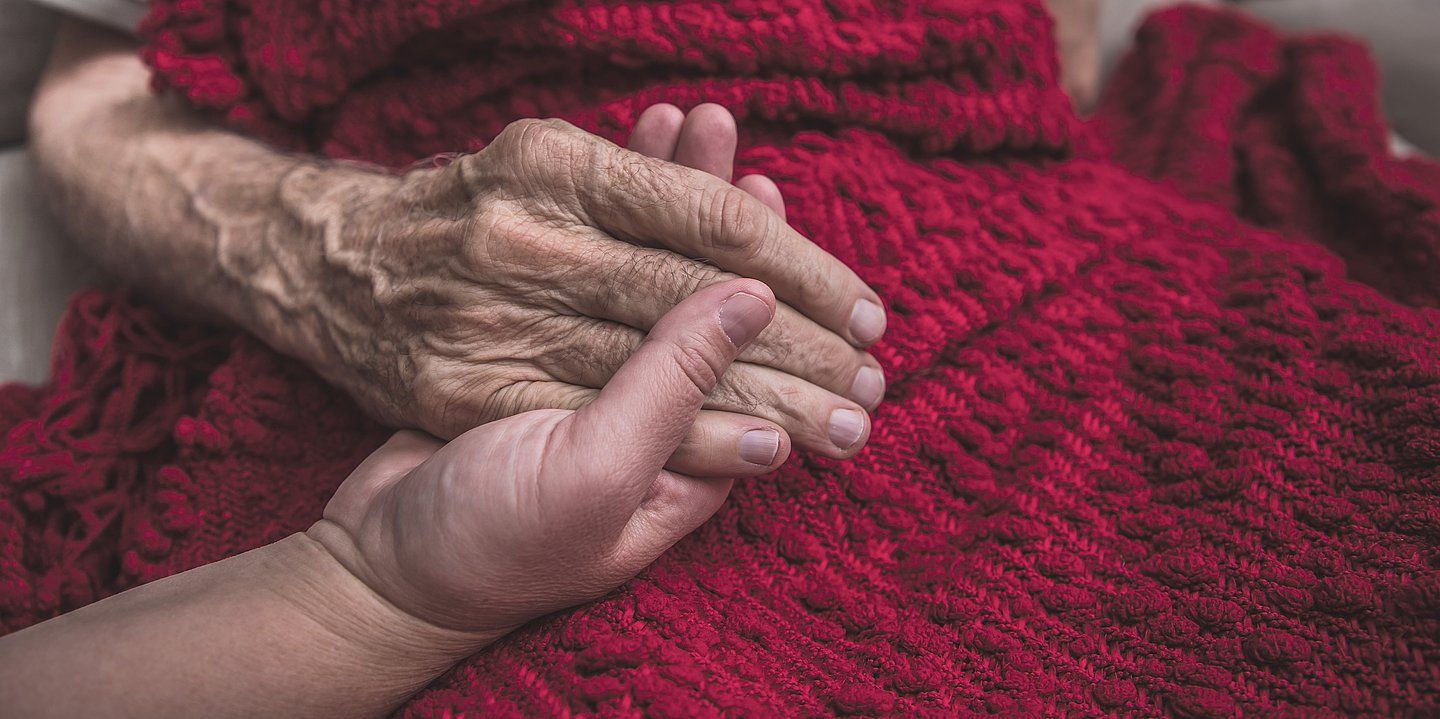 Foto: Eine jungaussehende Hand hält eine ältere aussehende Hand, die Hände liegen auf einer dunkelroten Wolldecke.