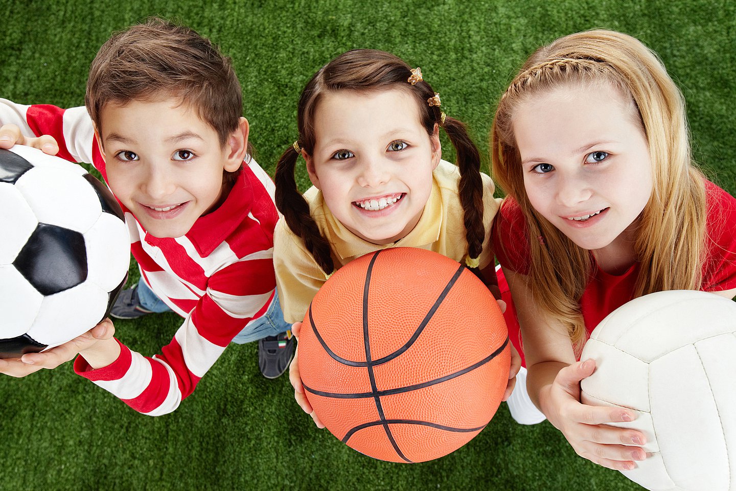 Foto von drei Kindern, die jeweils einen Ball halten, Fußball, Basketball, Handball, und lächelnd zum Betrachter nach oben schauen.