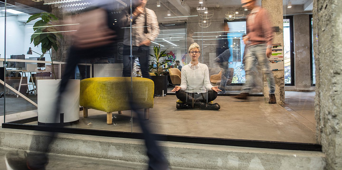 Foto einer Frau, die in einem modernen Büro meditiert. Um sie herum herrscht Hektik.