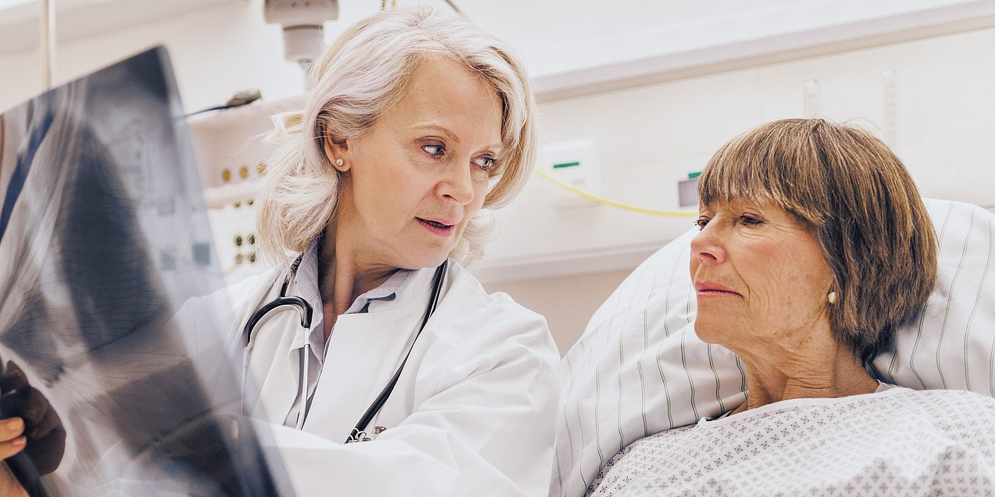 Symbolbild einer Ärztin, die einer Frau in einem Krankenhausbett ein Röntgenbild erklärt