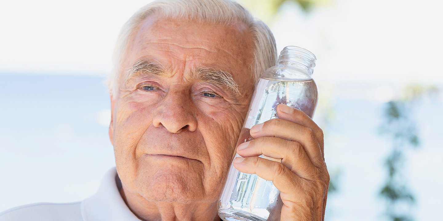 Foto: Ein älterer Mann hält sich eine Wasserflasche an die Wange.