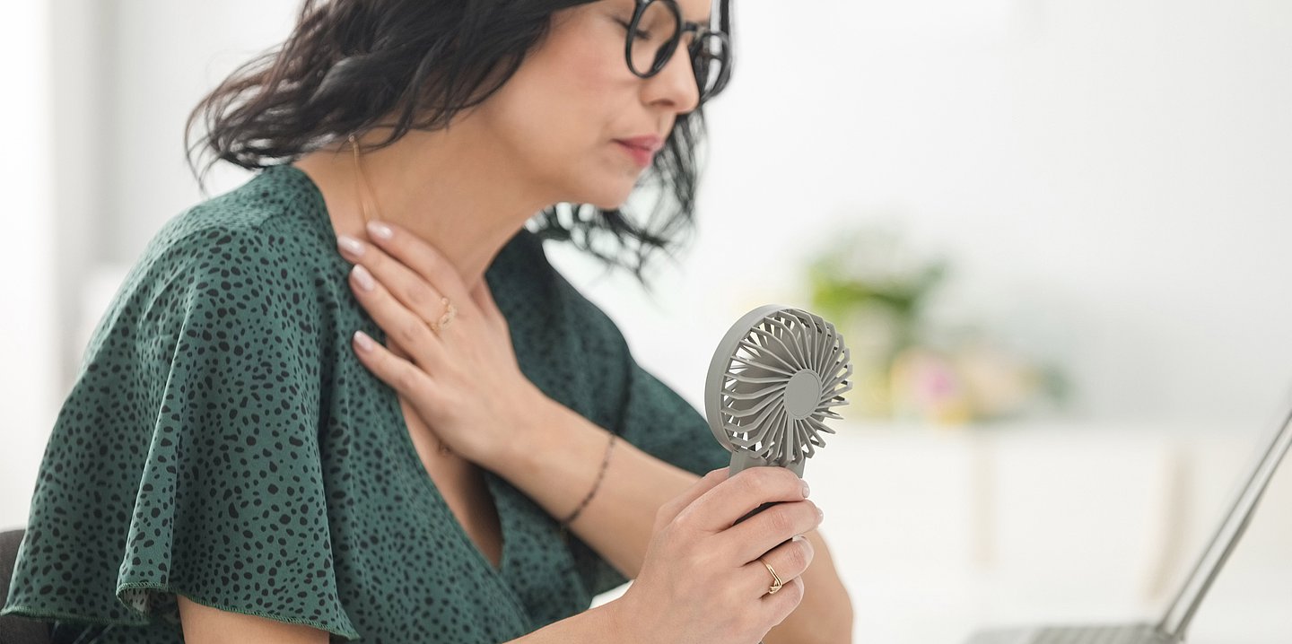 Foto einer Frau im mittleren Alter, die im Büro sitzt und sich schwitzend einen kleinen Ventilator vor den Körper hält