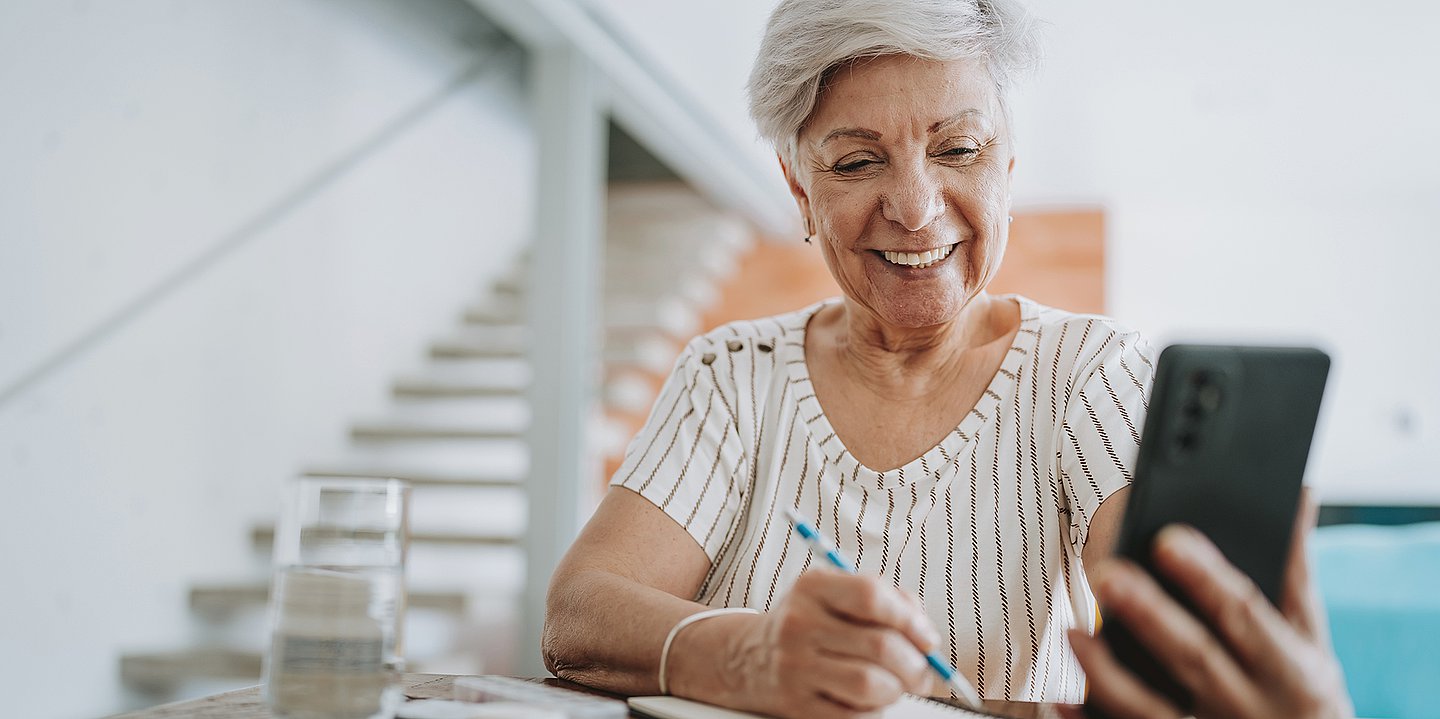 Foto: Eine ältere Dame hält lächeln ihr Smartphone in der Hand und blickt darauf, während sie gleichzeitig einen Stift in der Hand hält.