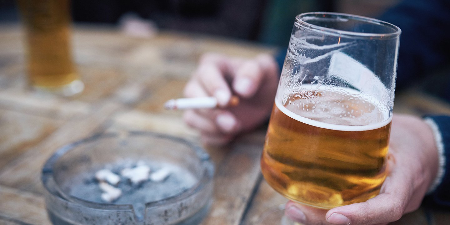 Foto: Eine Hand hält ein halb gefülltes Bierglas und hält eine Zigarette, daneben steht ein Aschenbecher.