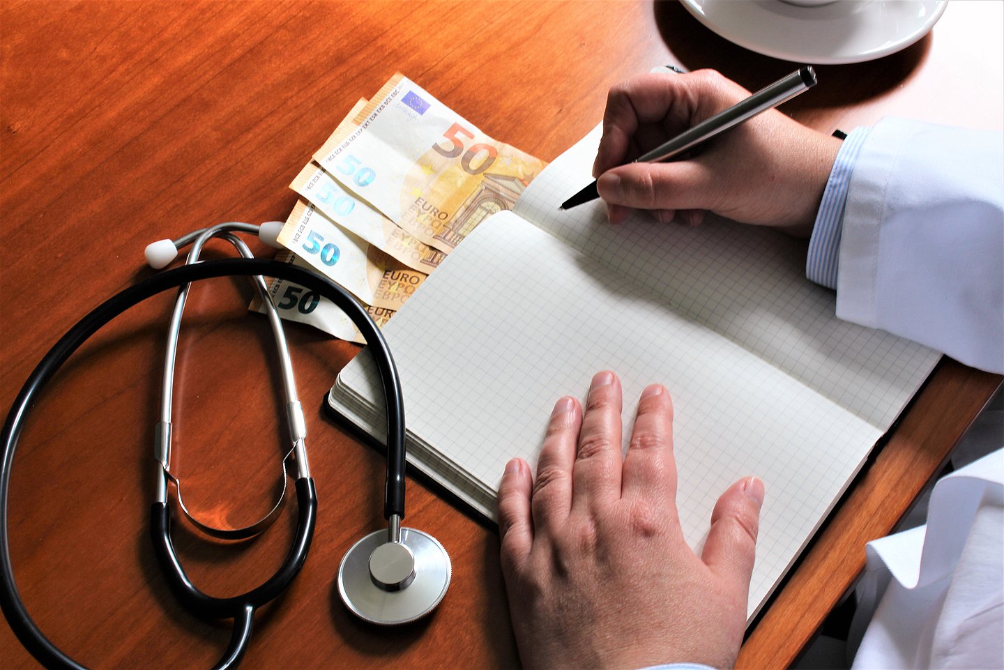 Das Foto zeigt einen Arzt, der an seinem Schreibtisch sitzend etwas in sein Notizbuch schreibt. Daneben lieben vier 50 Euro-Scheine.