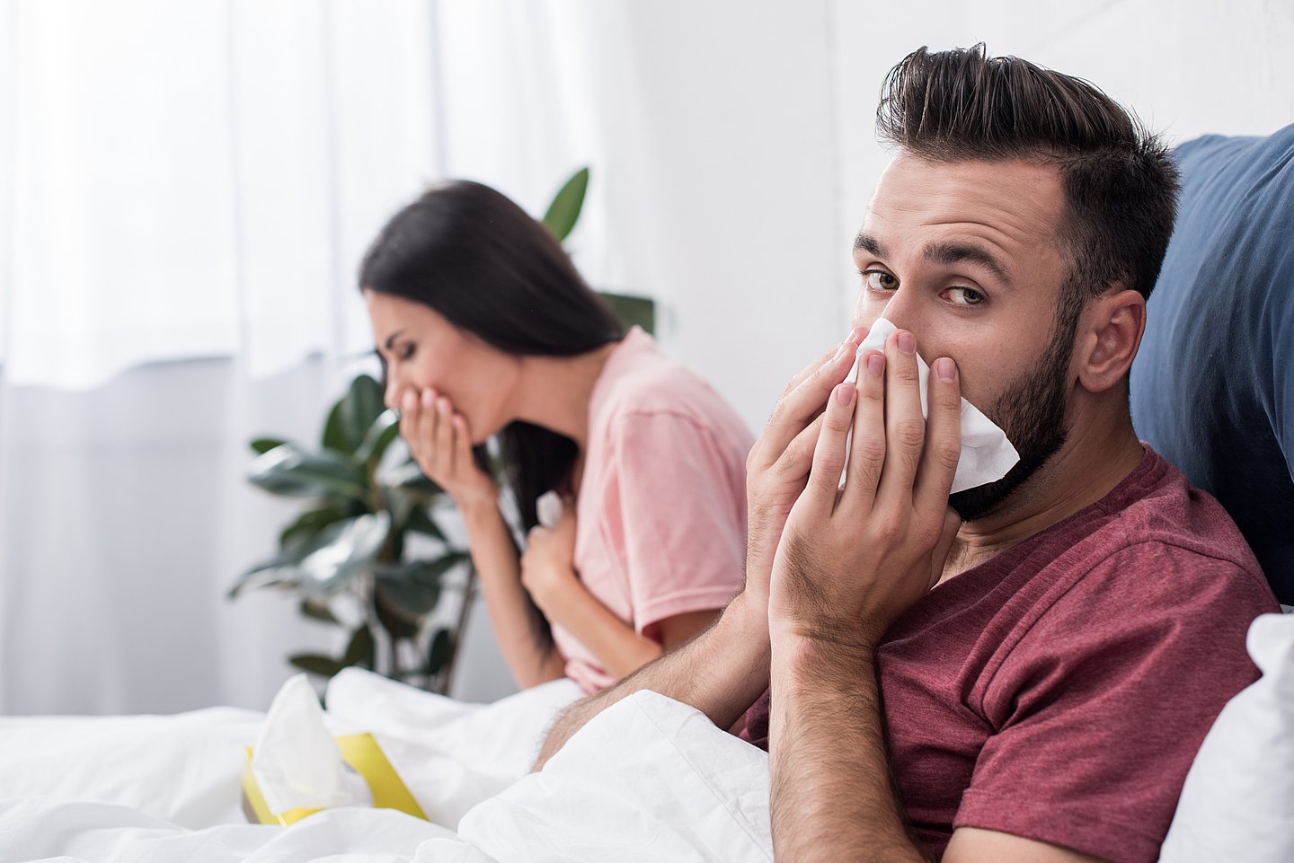 Foto zeigt eine Frau und ein Mann krank im Bett, sie hustet, er schnieft in ein Taschentuch und blickt in die Kamera.