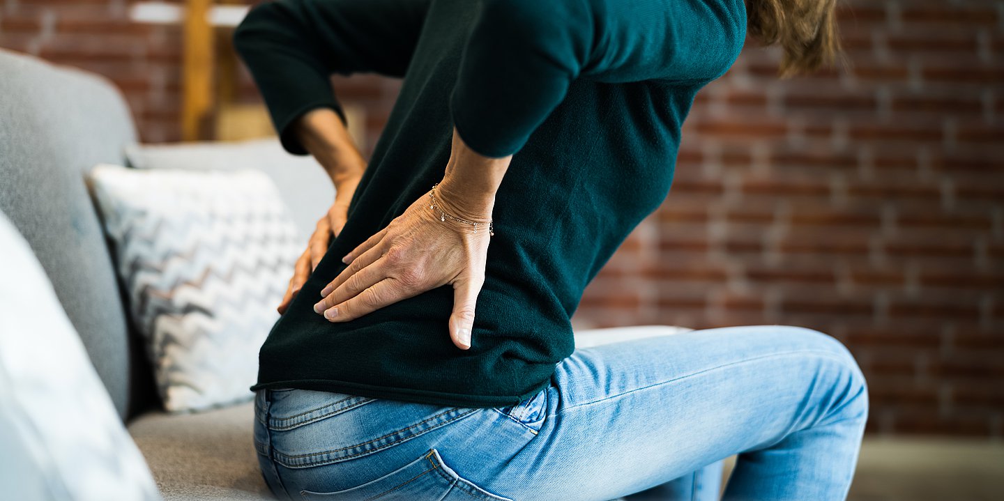 Eine Frau sitzt auf der Couch und hält sich ihren schmerzenden Rücken.