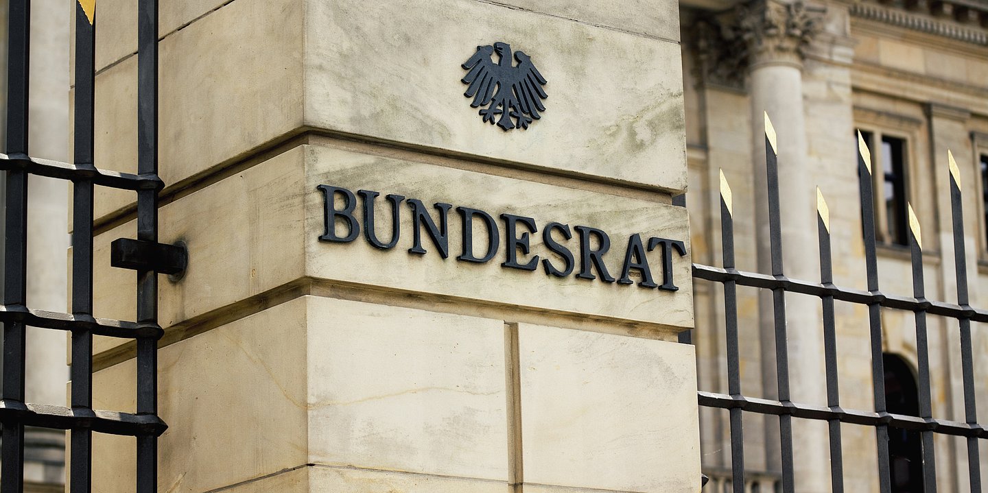 Das Foto zeigt den Eingangsbreich des Bundesrates in Berlin in Nahaufnahme. Zu sehen ist auf einer Säule der Schriftzug BUndesrat in Metalllettern, darüber der Bundesadler. Rechts und links säumt ein Metallzaun mit goldfabrenen Spitzen die Säule.