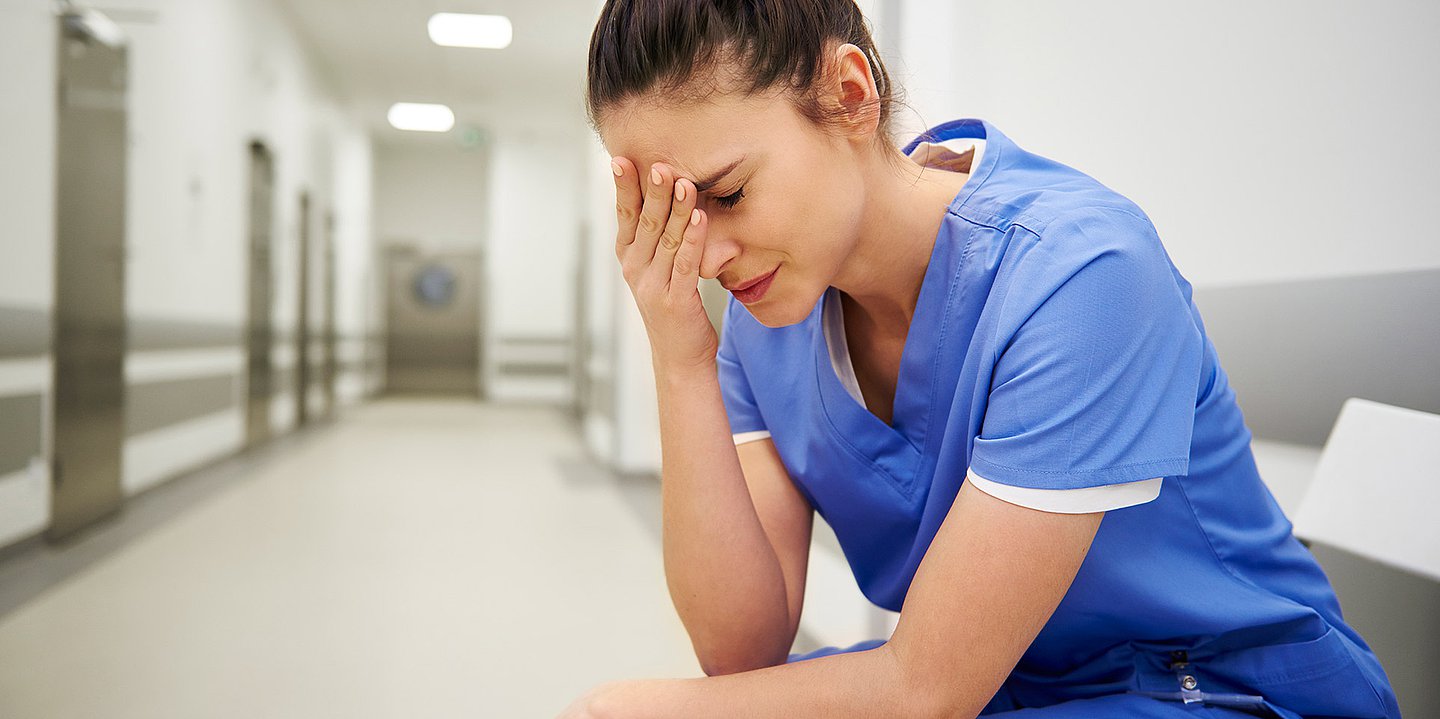 Foto: Eine Frau in blauer Pflegetracht sitzt auf einem Krankenhausflur und hält sich die Hand vor das Gesicht.