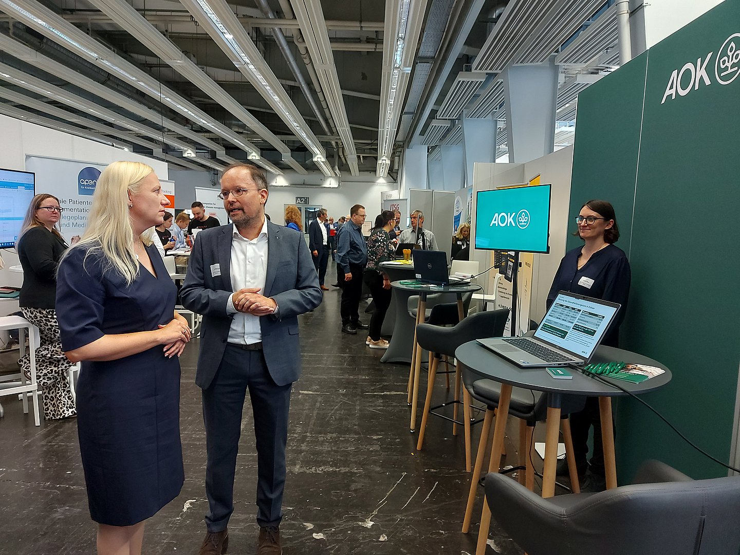 Das Bild zeigt eine blonde Frau in einem blauen Kleid (links) im Gespräch mit einem graumelierten Mann mit hoher Stirn in einer Art Messehalle. Rechts sieht man einen LED-Bildschirm mit dem AOK-Logo sowie mehrere Stehtische und Hocker aufgereiht.