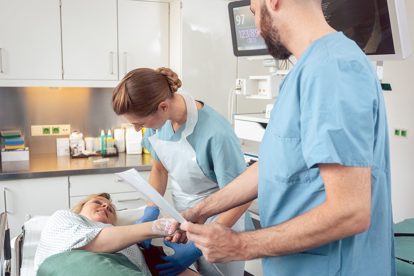 Eine weibliche Patientin liegt auf der Liege eines Behandlungsraums für eine Darmspiegelung. Eine Assistentin legt ihr eine Blutdruckmanschette um den linken Oberarm an und der behandelnde Arzt begrüßt die Patientin per Handschlag. An der Hand sieht man den bereits gelegten Zugang.