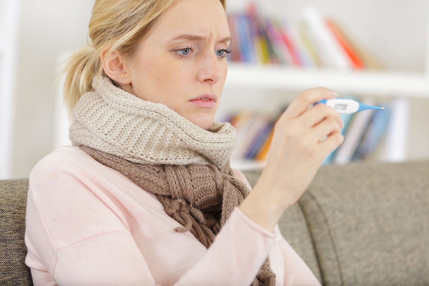 Eine Frau sitzt mit einem Schal um den Hals auf der Couch und schaut auf ein Fieberthermometer.