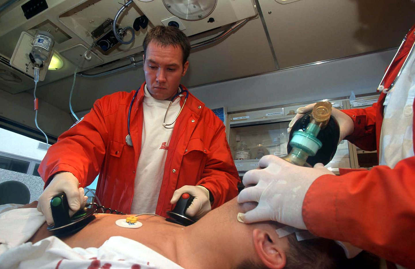 Das Bild zeigt zwei Rettungssanitäter im Rettungswagen bei der Wiederbelebung eines Patienten mit Defibrilator und Sauerstoffsack