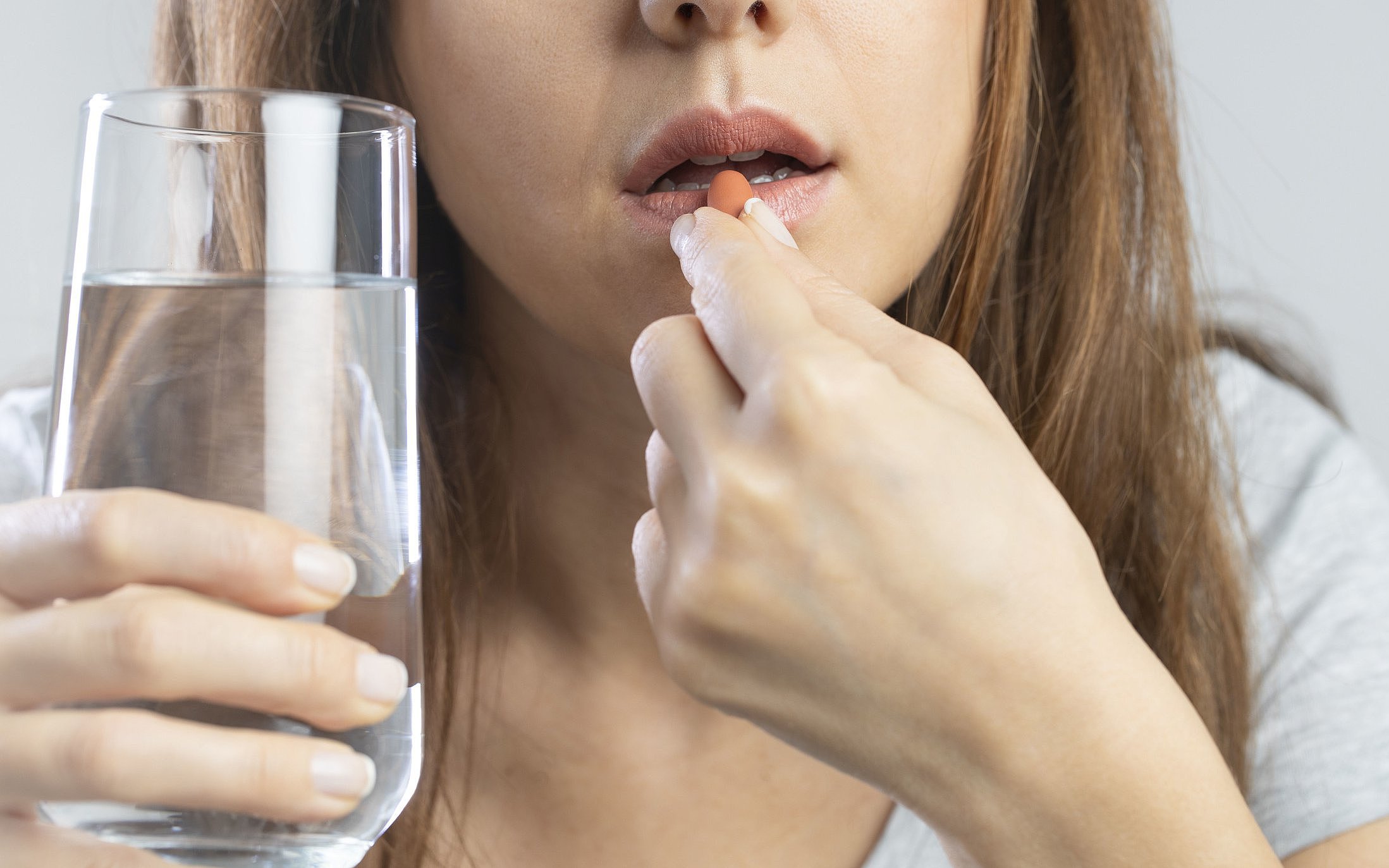 Auf dem Bild ist eine Frau mit einem Glas Wasser in der Hand zu sehen, die eine Tablette einnimmt. 