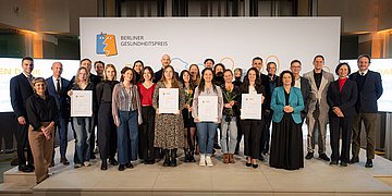 Die Gewinnerinnen und Gewinner des Berliner Gesundheitspreises stehen auf der Bühne zusammen mit Dr. Carola Reimann, Vorstandsvorsitzende des AOK Bundesverbandes und Dr. Peter Bobbert, dem Präsidenten der Ärztekammer Berlin und halten ihre Urkunden in den Händen.