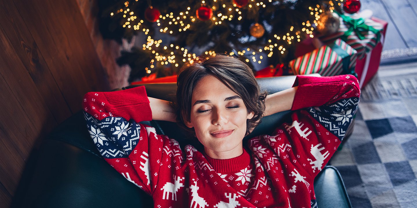 Foto: Eine Frau im Weihnachtspullover liegt entspannt mit geschlossenen Augen auf der Couch. 