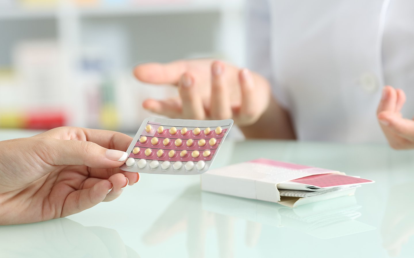 Foto zeigt einen Tisch auf dem eine Packung Antibabypillen liegt. Auf der Seite des Betrachters hält eine Hand einen Blister mit den Pillen, auf der anderen Seite des Tisches sieht man den Ausschnitt einer Person in weißem Kittel, deren Hände gestikulieren. Person 