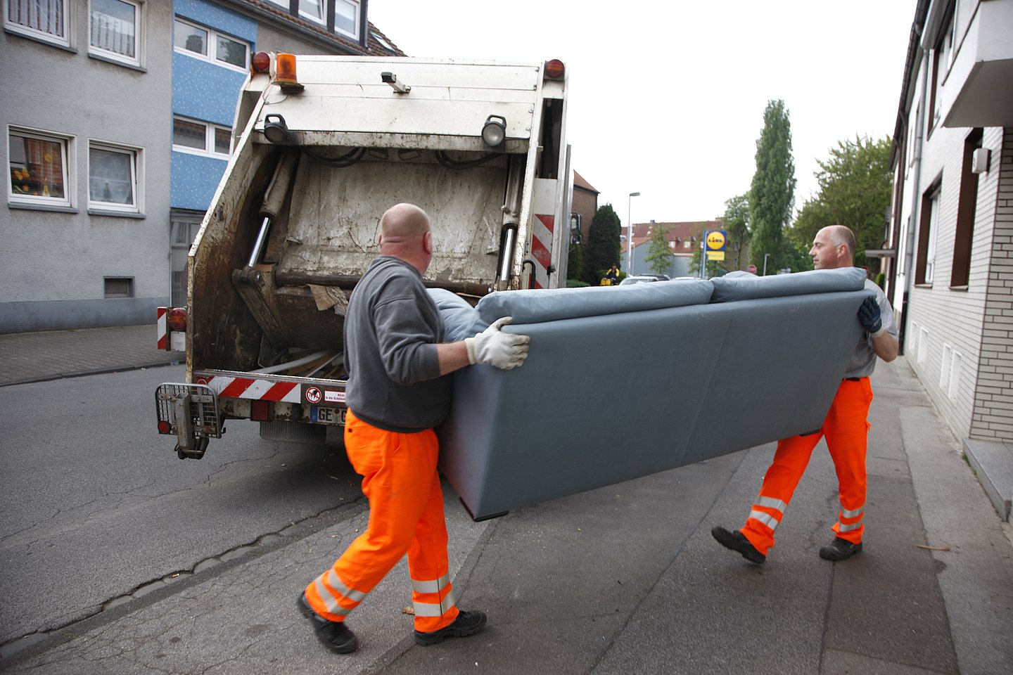 Das Foto zeigt zwei Müllweker in klassischer orangefarbener Arbeitskleidung. Sie tragen ein ausgedientes hellblaues Sofa zum Müllwagen, der im Hintergrund steht.