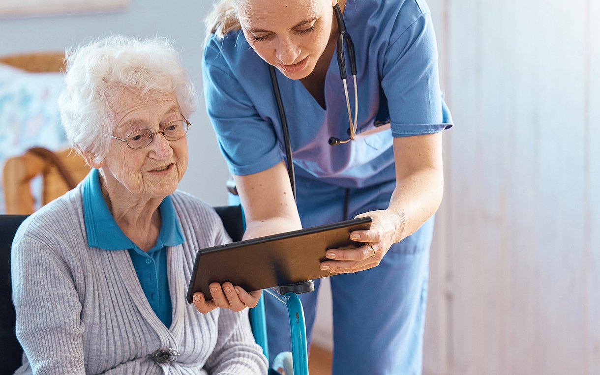 Foto einer Pflegerin, die sich mit Tablet zu einer alten Frau im Rollstuhl herunterbeugt und ihr etwas erklärt.