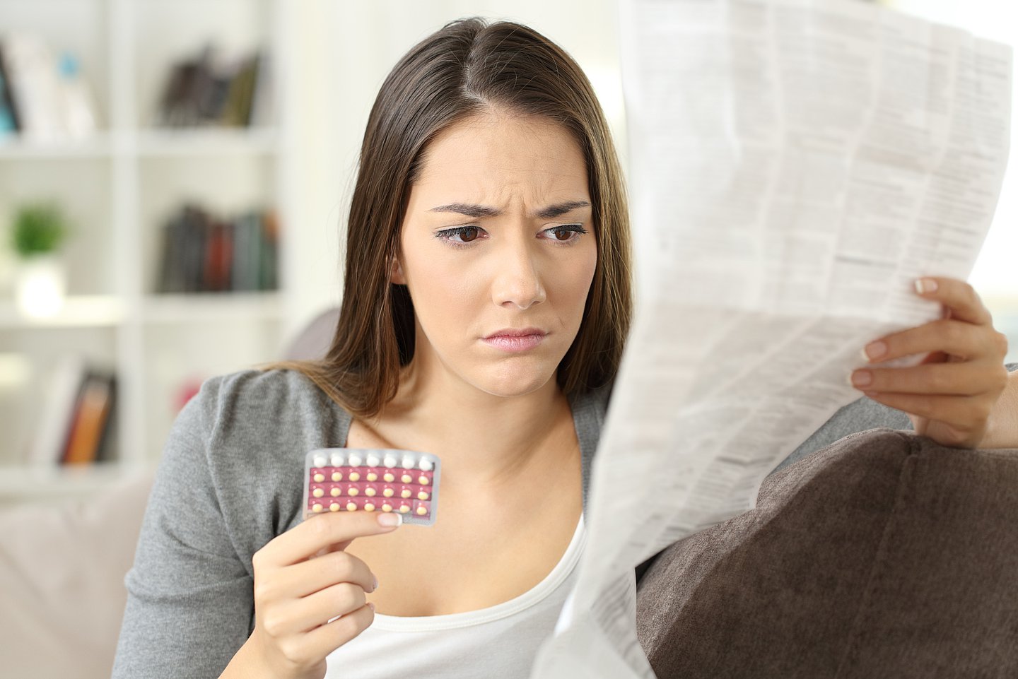 Foto zeigt eine auf einem Sofa sitzende Frau, die in der einen Hand eine Packung mit Antibabypillen in der Hand hält, in der anderen den Beipackzettel dazu, den sie mit skeptischem Blick durchliest.