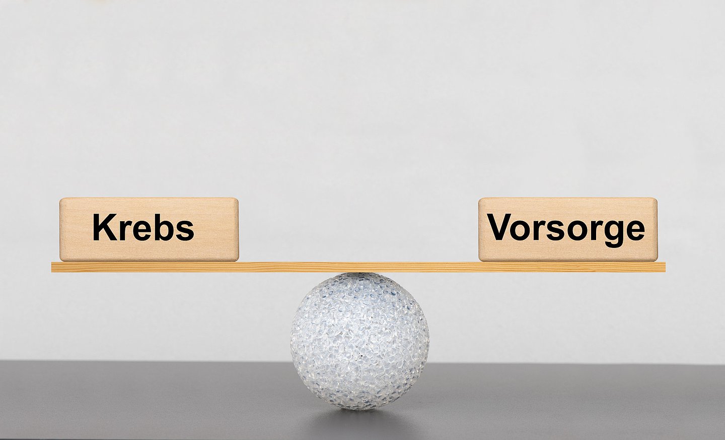 Mit einem kleinen Ball und einem Holzbrett mit den Gewichten 'Krebs' und 'Vorsorge' wird eine Waage dargestellt, die im Gleichgewicht ist.