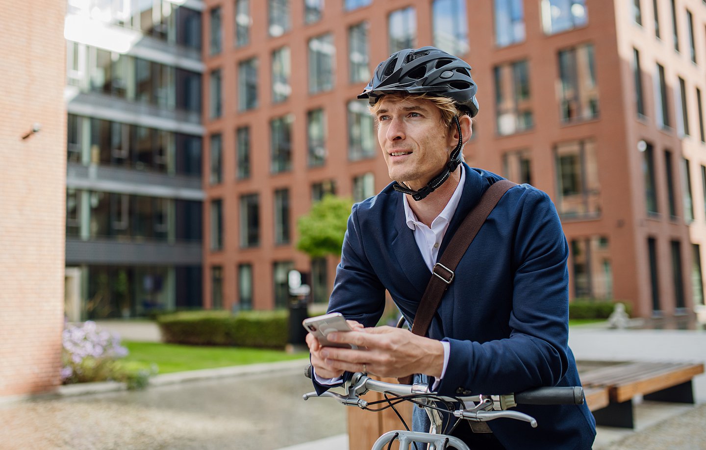 Ein junger Mann steht mit seinem Fahrrad vor weinem Bürogebäude. Er stützt sich sich auf den Lenker und hält ein Smartphone in seinen Händen.Er trägt einen Fahrradhelm, ein dunkelblaues Sakko, darunter ein blasslaues Oberhemd. Um den Rumpf liegt der braune Lederriemen seiner Aktentasche. Im Hintergrund sind vor dem Bürogebäude Bäume, eine Hecke und ein Rasen zu sehen.