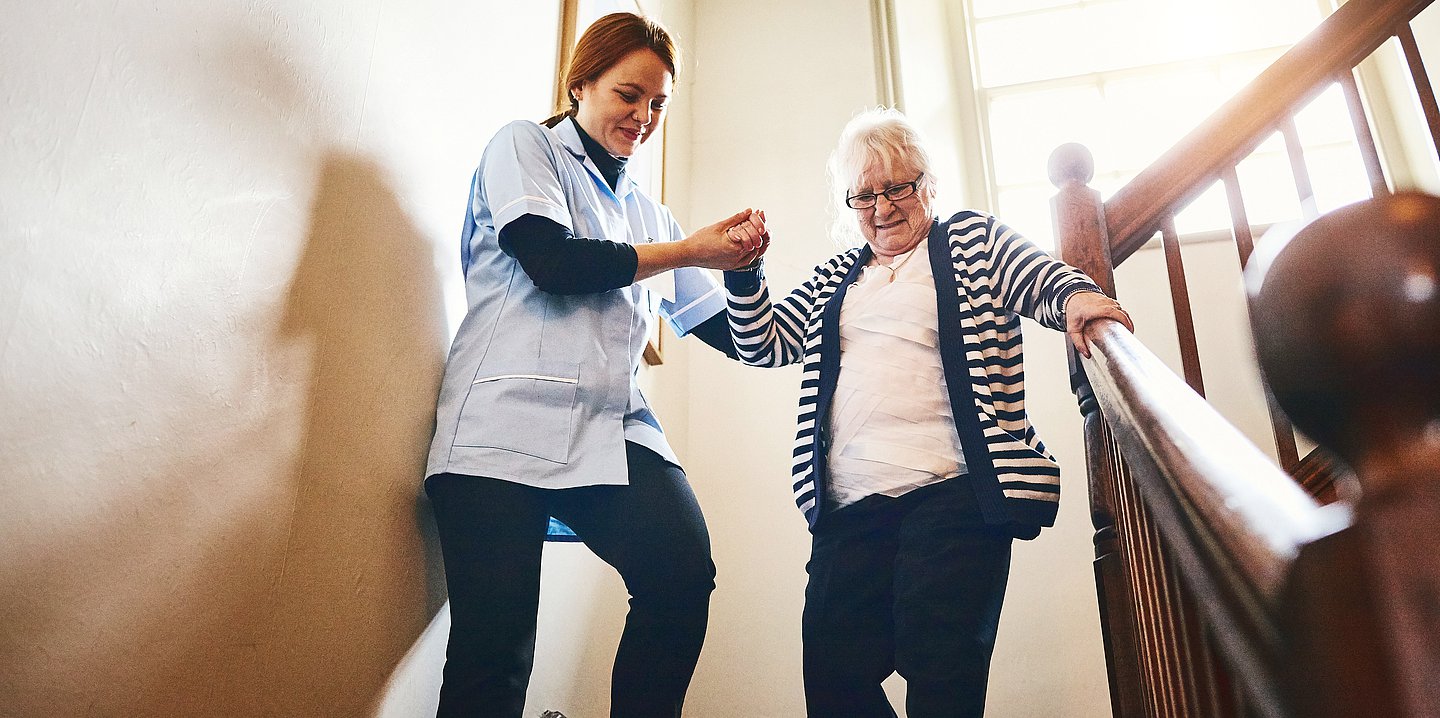 Foto: Eine jüngere Dame in Pflegekleidung hilft einer älteren Dame die Treppe hinunterzusteigen.