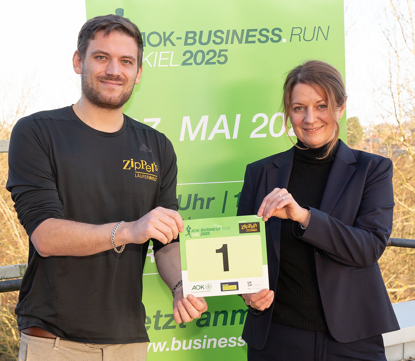 Ein junger Mann und eine Frau stehen vor einem Roll-up, auf dem die Daten des Laufs vermerkt sind und halten beide gemeinsam die Startnummer "1" in der Hand.