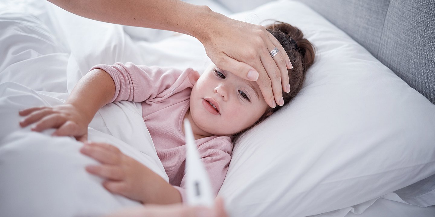 Ein Kleinkind liegt im Bett, die Mutter hält eine Hand auf den Kopf des Kindes und ein Fieberthermometer in der anderen Hand.