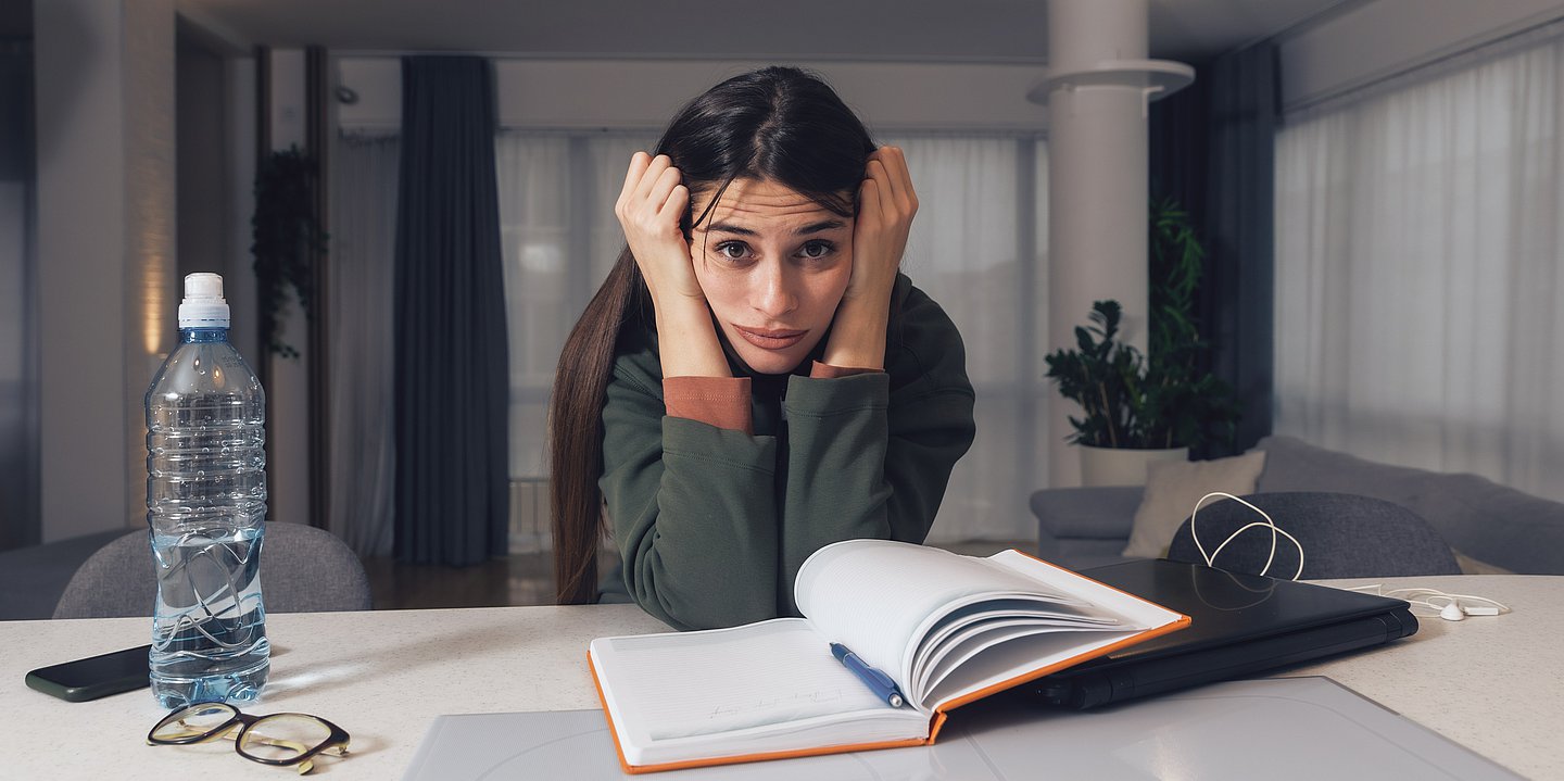 Foto: Ein Frau sitzt am Tisch über einem aufgeschlagenem Buch und guckt frustriert.