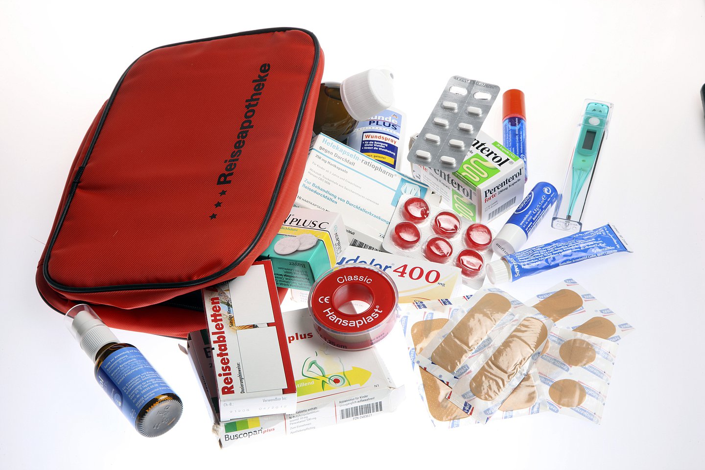 Bild zeigt ein rotes Täschechen mit der Aufschrift "Reiseapotheke". Drumherum verscheiedene Medikamente un dMediziprodukte wie Tabeltte, Nasenspray, Pflaster  und Fieberthermometer.