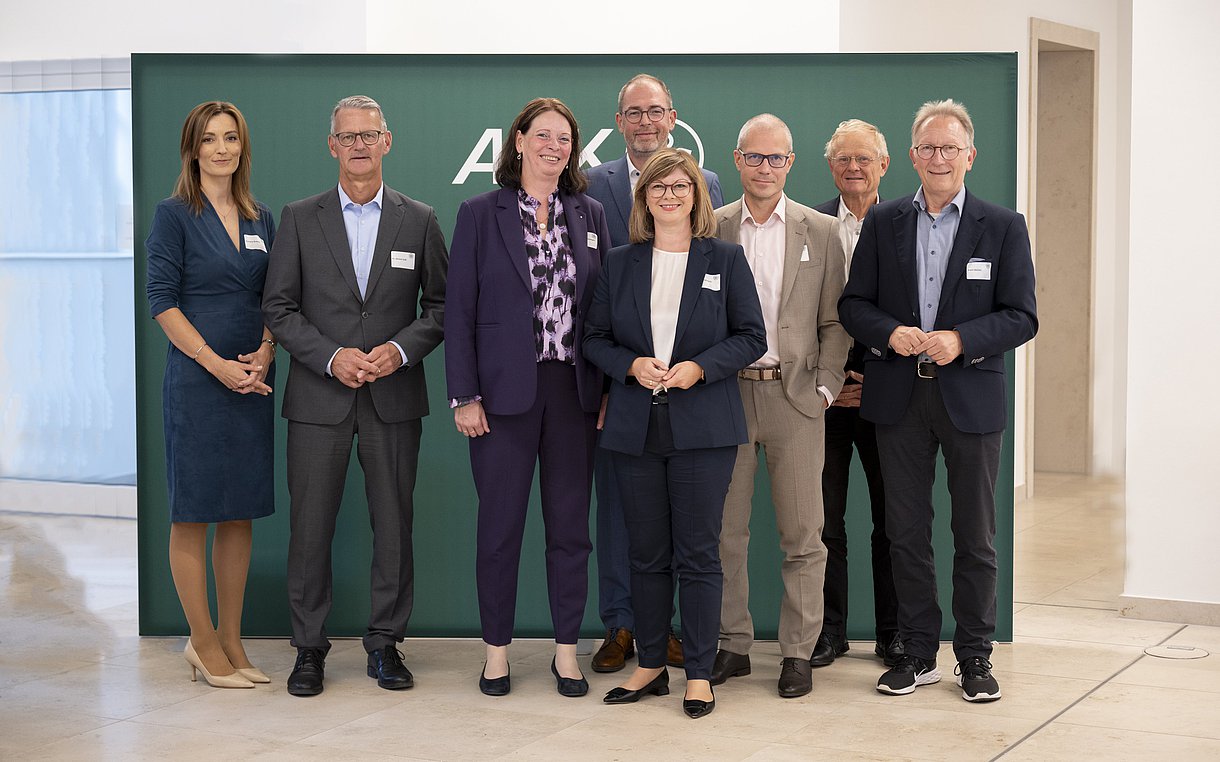 Das Foto zeigt ein Gruppenbild mit insgesamt acht Personen (drei Frauen, fünf Männer). Auf der dunkelgrünen Wand im Hintergrund ist schemenhaft das AOK-Logo zu sehen.