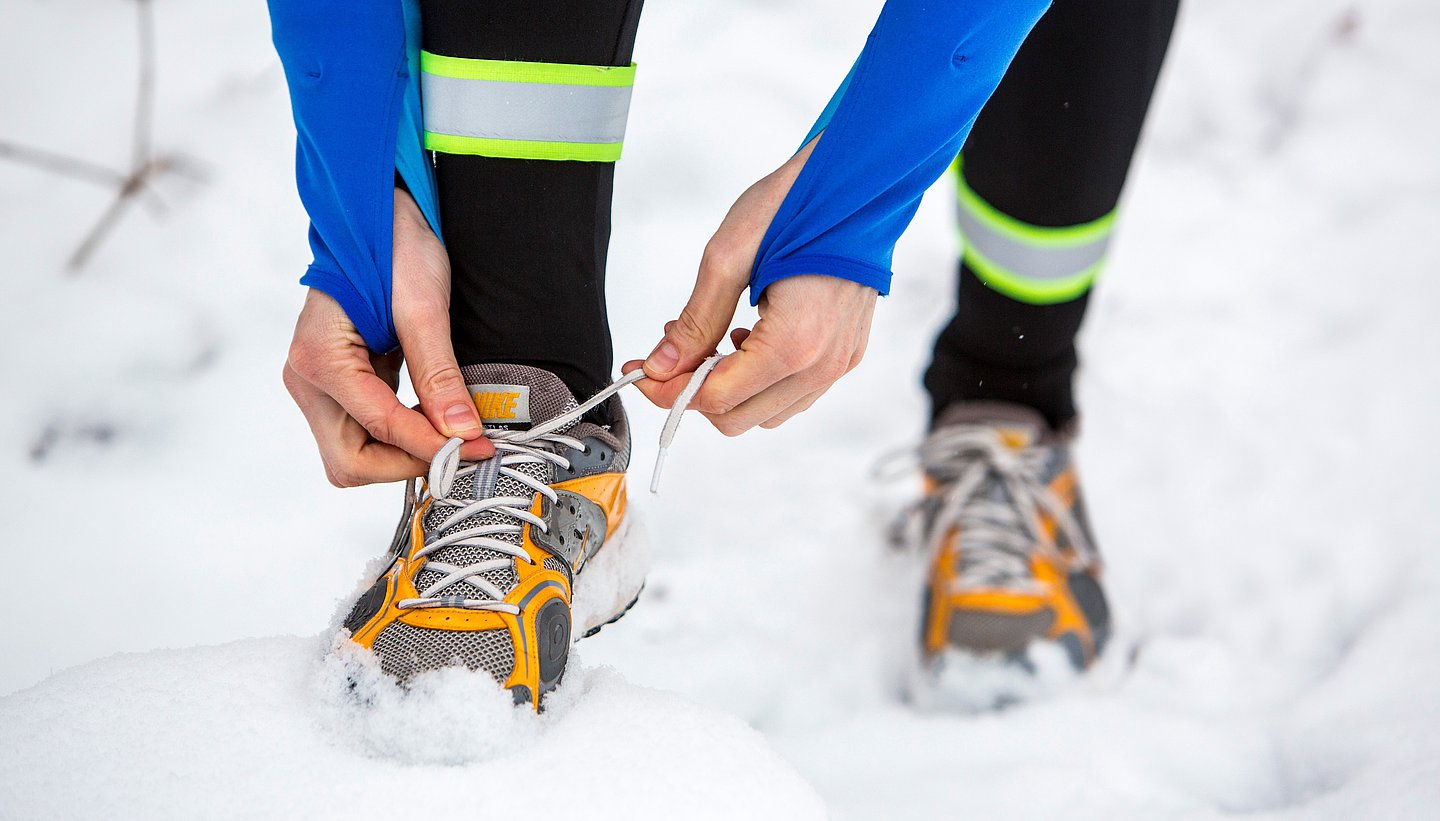 Das Bild zeigt Füße und Beine unterhalb des Knies auf Echneeunterlage und zwei Hände, die den rechten Joggingschuh schnüren.